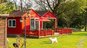 Une cabane en bois rouge avec une terrasse, une pelouse et un petit chien.