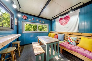 Une pièce avec une table en bois, deux tabourets, un banc agrémenté de coussins colorés, une fenêtre donnant sur un paysage verdoyant, et un mur décoré d’un motif en forme de cœur et d’une inscription.