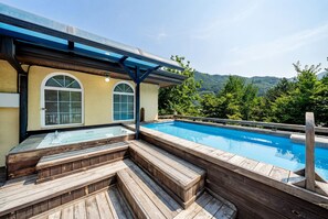 A wooden deck with a hot tub and a swimming pool, surrounded by a house with arched windows and a view of a forested hillside. - Gapyeong Ruba Pension (Gapyeong)