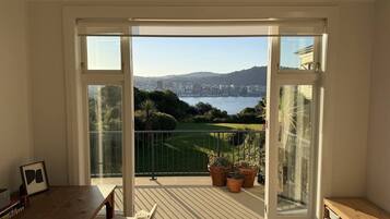 A balcony with a view of a city and water, featuring a wooden table, chairs, and potted plants.