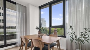 A dining area with a wooden table and chairs, large windows with curtains, and potted plants. - Brand New Apartment in Albert Park with Pool & Gym (Melbourne)