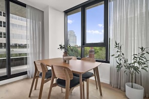 Un comedor con una mesa y sillas de madera, ventanas grandes con cortinas y plantas en macetas.