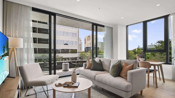 A modern living room with a grey sofa, a coffee table, and a TV. There is a balcony with a view of buildings and greenery. - Brand New Apartment in Albert Park with Pool & Gym (Melbourne)