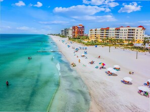 Uma praia com águas turquesa cristalinas, areia branca e guarda-sóis coloridos. Edifícios com telhados vermelhos ao fundo.