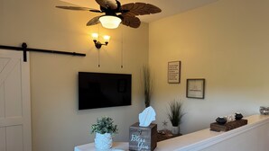 Un salon doté d’un ventilateur de plafond, d’un téléviseur fixé au mur, d’une console blanche agrémentée d’objets décoratifs et d’un luminaire fixé au mur.