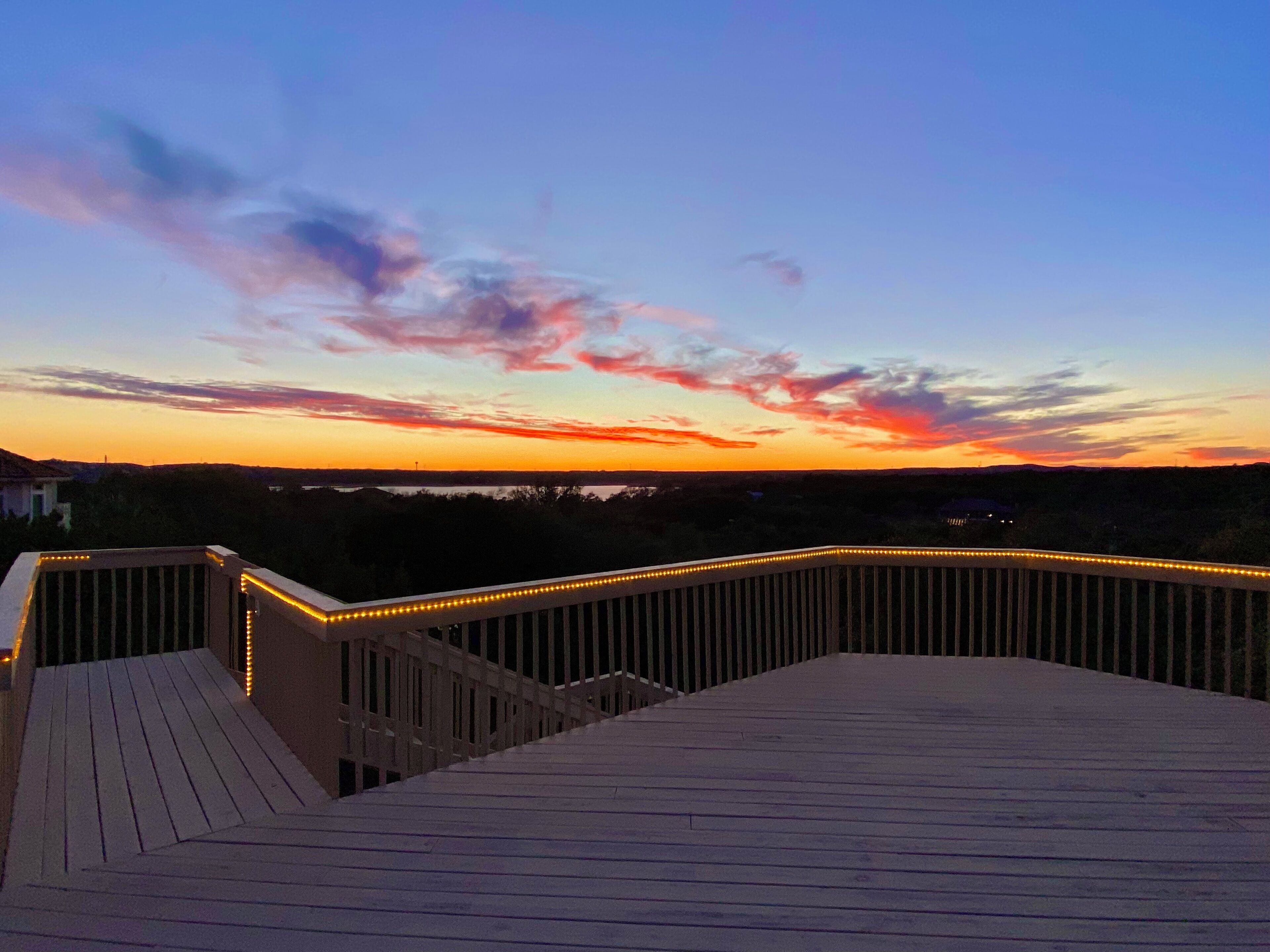 A wooden deck with railings and string lights, overlooking a landscape with distant buildings and a sunset sky.