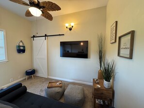 Uma sala de estar com sofá, pufes, televisão fixada na parede, ventilador de teto e porta deslizante.