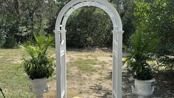 A white arched structure with a stone pathway leading through it, surrounded by greenery and potted plants.