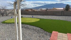 A well-maintained outdoor area with a green lawn, two blue flags, and a view of mountains in the background. - Newly Remodeled Mountain Escape Near Casino + Pool Table + Putting Green (Rio Rancho)