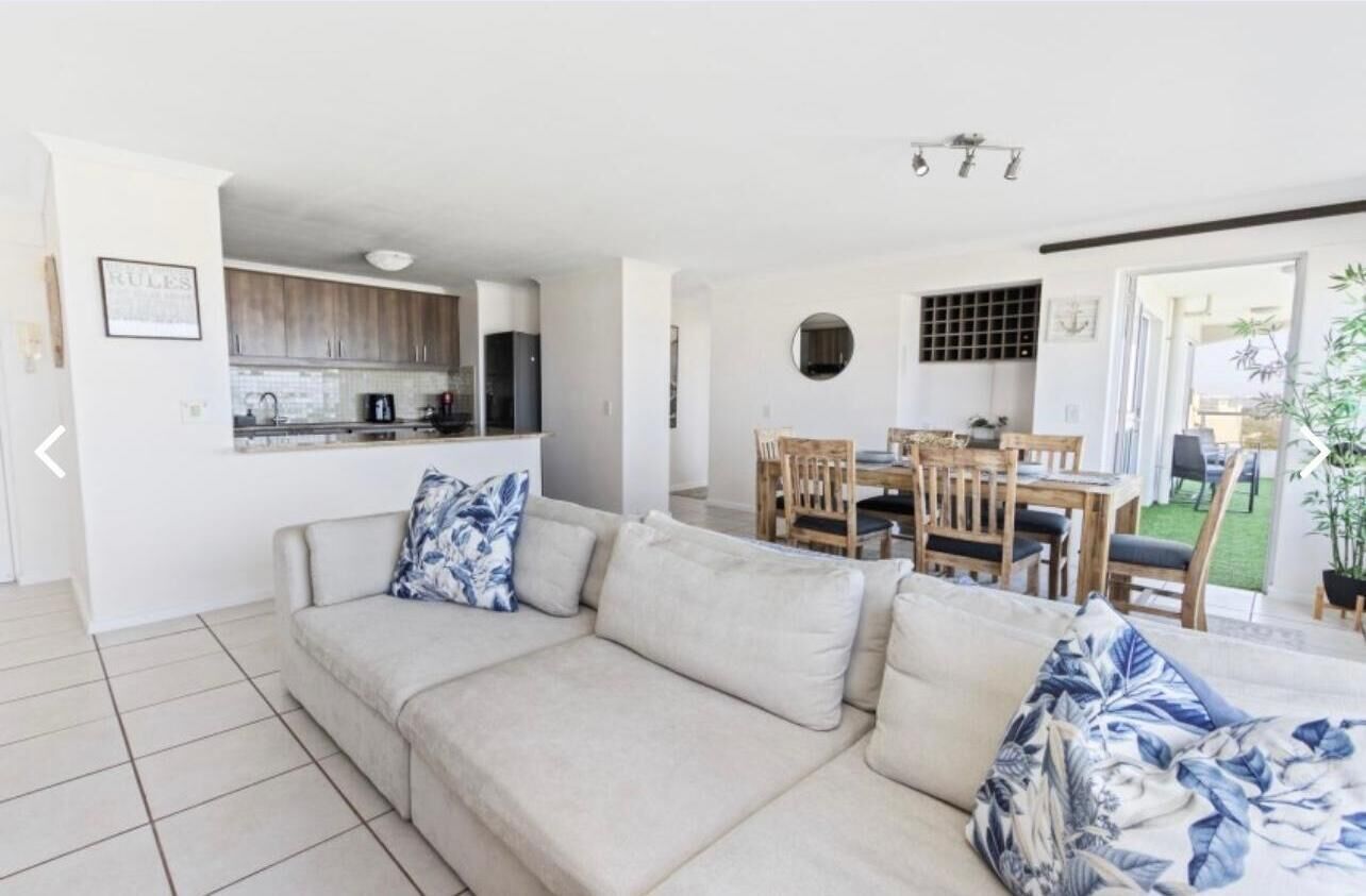 Un salon avec un canapé beige, une table à manger avec des chaises, et un coin cuisine à l’arrière-plan.