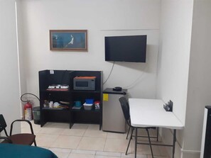 Une petite pièce propre, avec une table blanche, une chaise noire, un téléviseur fixé au mur, une étagère contenant divers objets et un extincteur.