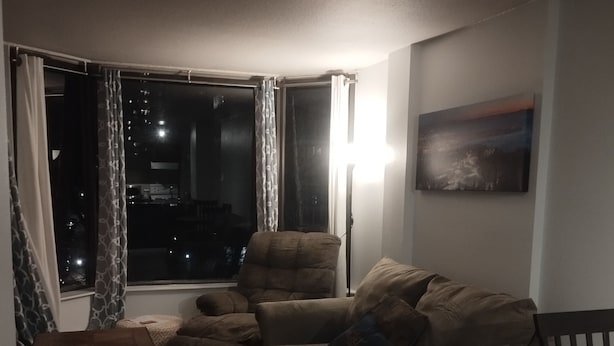 Ein Wohnzimmer mit einer Couch, einem kleinen Tisch und einer Lampe. Ein Fenster mit Vorhängen und ein Gemälde an der Wand.