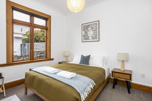 Une chambre avec une grande fenêtre, un lit recouvert d’une couverture verte, deux tables de chevet avec des lampes et un tableau encadré au mur.