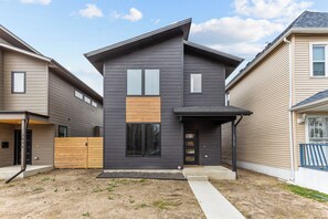 Une maison moderne à deux étages, dotée d’une façade sombre, d’un portail de garage visible et d’un petit jardin à l’avant, avec un chemin en béton.