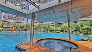 A modern hotel pool area with a hot tub, wooden deck, and a view of high-rise buildings. - SedaYu By SanLiving - 1BR - PIK (Jakarta)