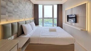 A modern hotel room with a large bed, a flat-screen TV mounted on the wall, and a view of greenery through the window. - SedaYu By SanLiving - 1BR - PIK (Jakarta)