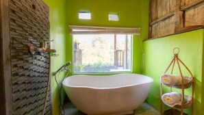 A bathroom with a freestanding bathtub, a window with a view of greenery, and a wicker towel rack. - Room-deluxe Bungalow at Doni House (Ubud)