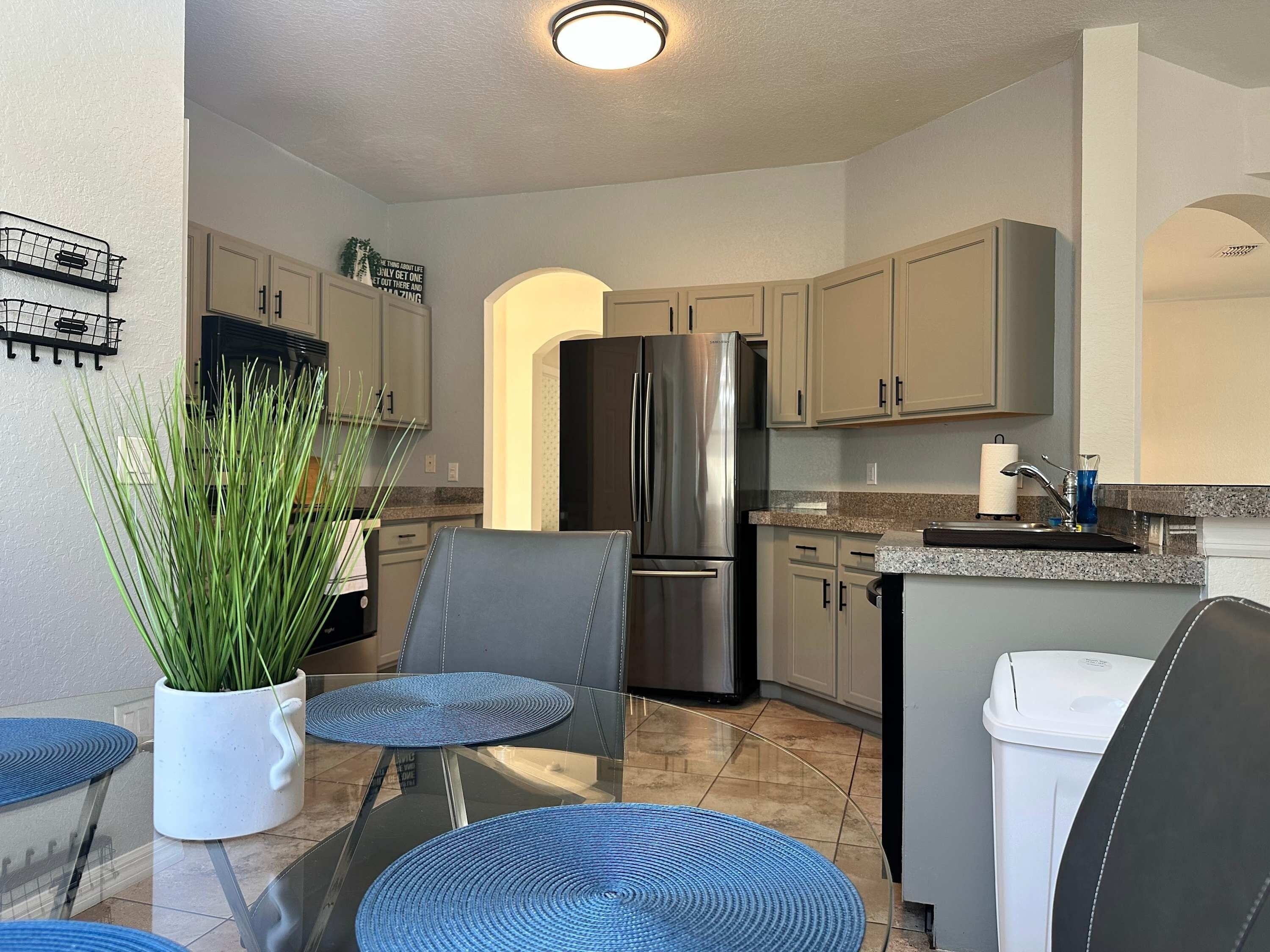 A compact kitchen with a stainless steel refrigerator, a sink, and a dining area with a chair and a potted plant.