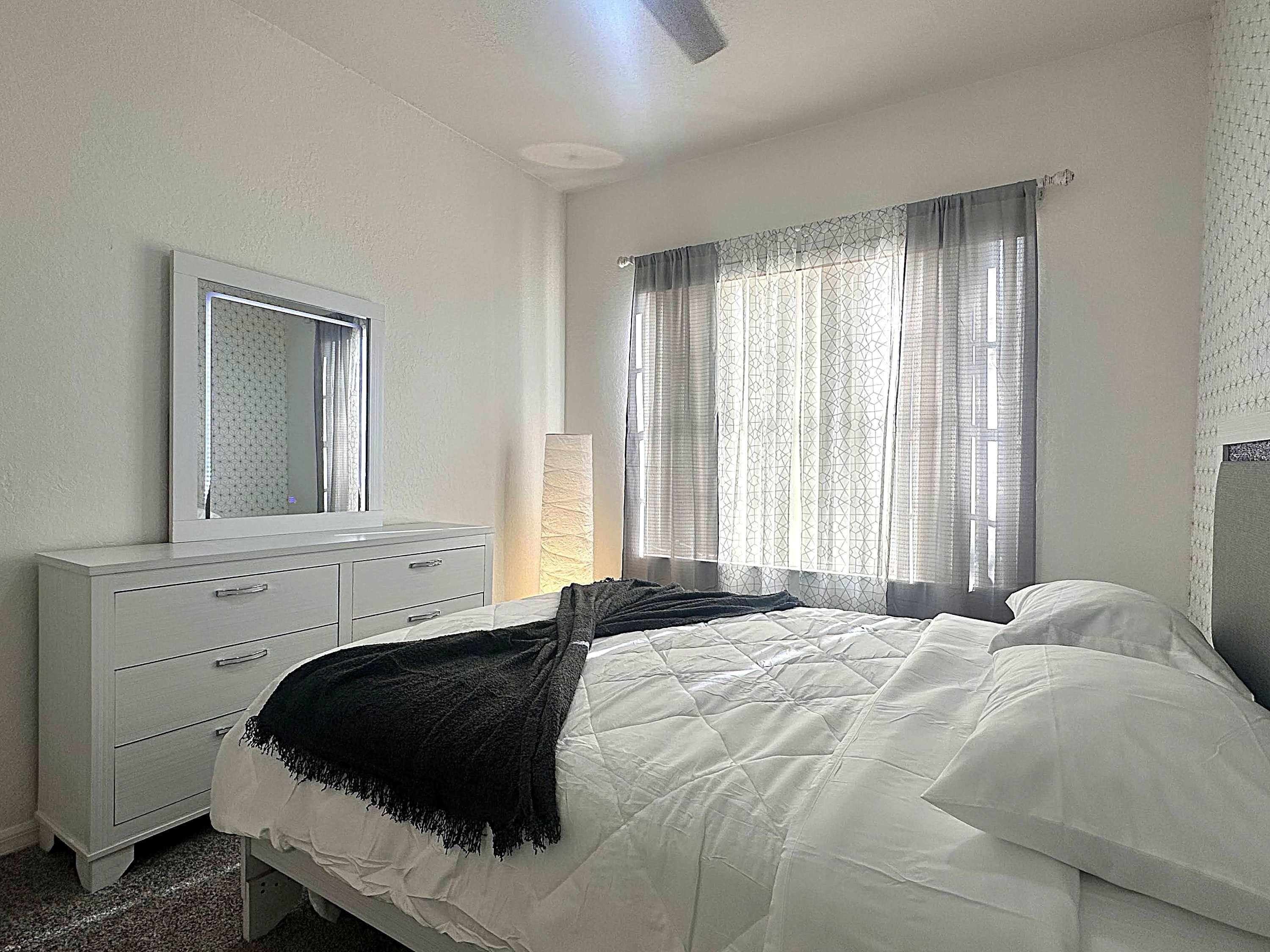 Une chambre à coucher avec un lit, une commode, un miroir et une fenêtre avec des rideaux.