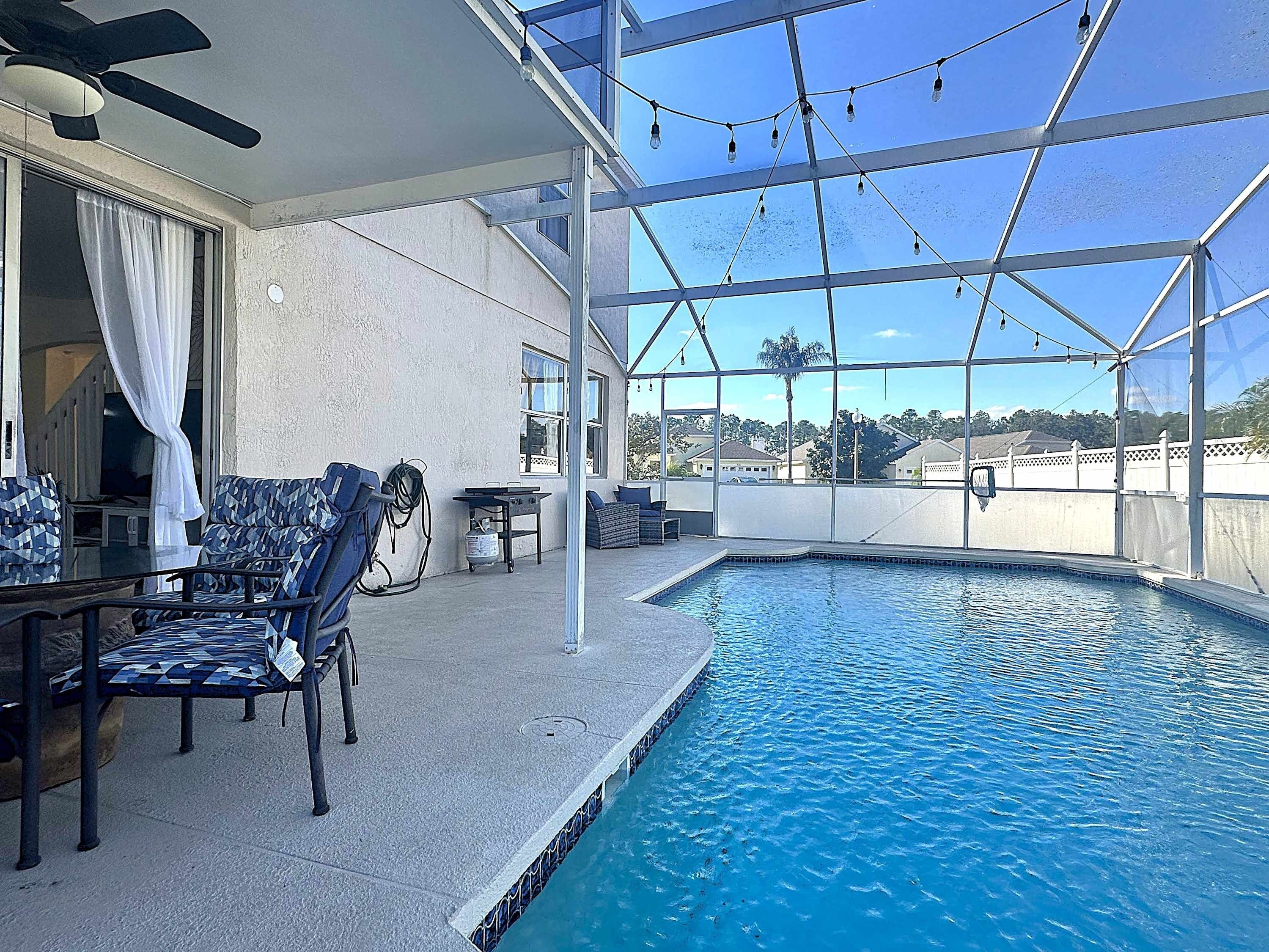 An indoor swimming pool with a ceiling fan, string lights, and a view of the surrounding neighborhood.