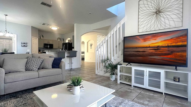 A modern living room with a sofa, coffee table, and TV. There is a kitchen in the background with white cabinets and a dining area with bar stools. - Family Villa 5 bedroom near disney world 5 Villa (Davenport)