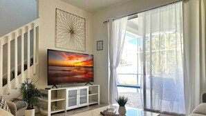 A living room with a large flat-screen TV on a white stand, a coffee table with books and a plant, a staircase with white railings, and a view of a pool through sheer curtains. - Family Villa 5 bedroom near disney world 5 Villa (Davenport)
