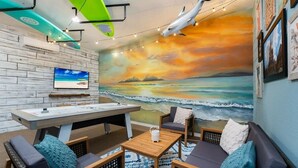 Habitación con un mural de playa, un televisor de pantalla plana, una mesa de madera y asientos con cojines.