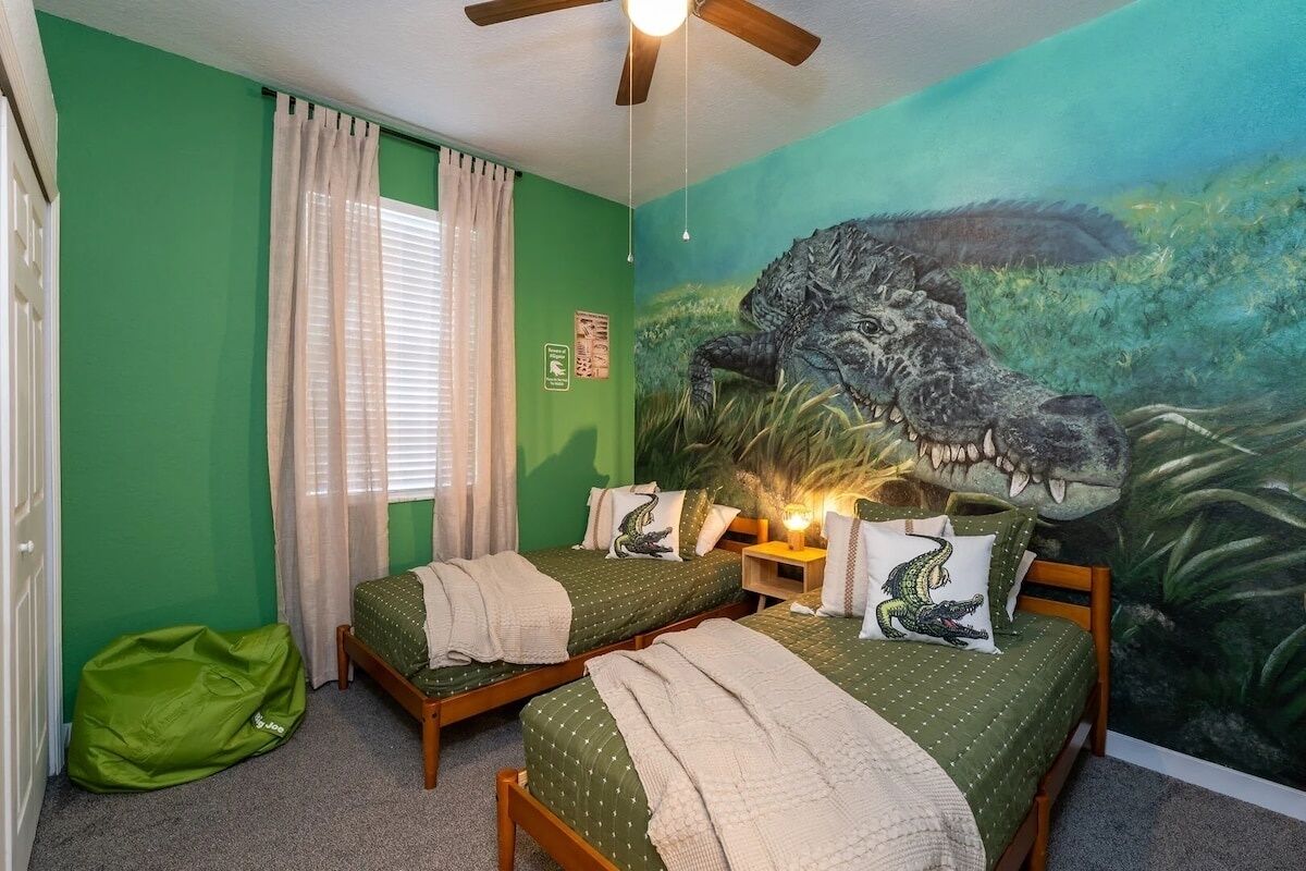 Een kamer met twee bedden, een plafondventilator, een groene wand, een muurschildering van een krokodil en een raam met gordijnen.