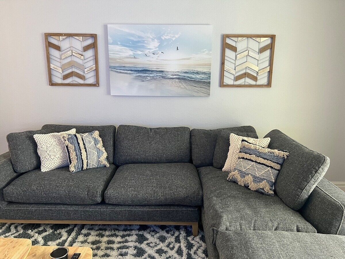 A grey sectional sofa with patterned cushions, a framed artwork of a beach scene, and a chevron-patterned wall decoration.