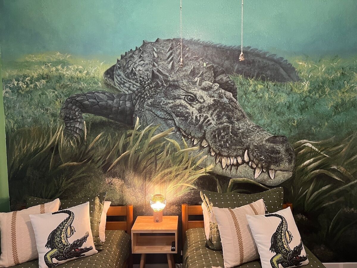 A bedroom with a mural of a crocodile, a bedside table with a lamp, and patterned pillows.