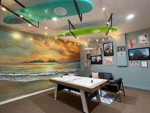 Ein Zimmer mit Strandwandgemälde, einem Tischfußballspiel und Surfbrettern, die von der Decke hängen.