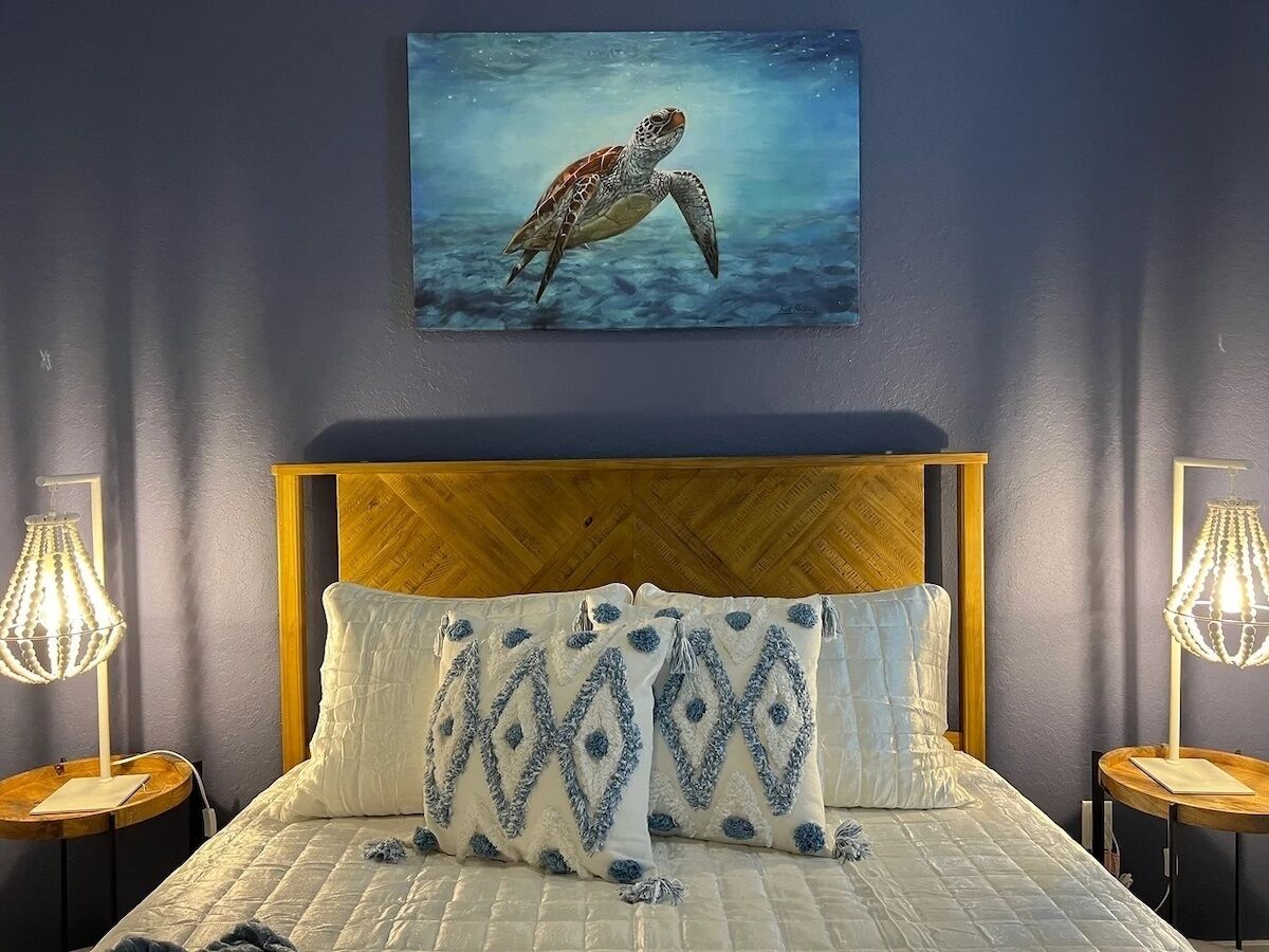Une chambre à coucher avec une tête de lit en bois, un lit agrémenté d’oreillers à motifs, un tableau représentant une tortue de mer et deux lampes de chevet.