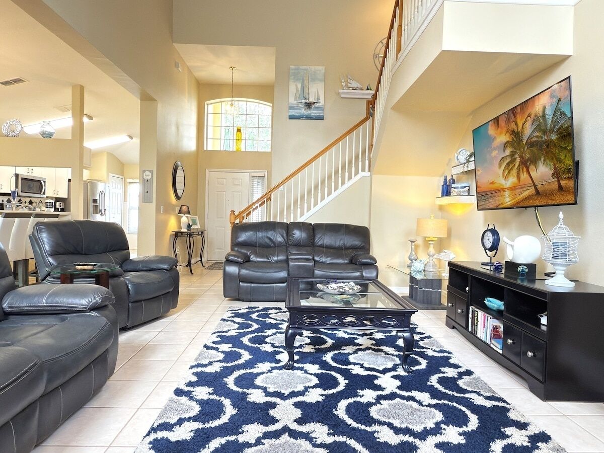 A spacious living room with a large black leather sofa set, a glass coffee table, and a patterned rug. A staircase with a wooden railing leads to an upper level.