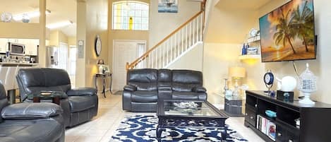 A spacious living room with a large black leather sofa set, a glass coffee table, and a patterned rug. A staircase with a wooden railing leads to an upper level.