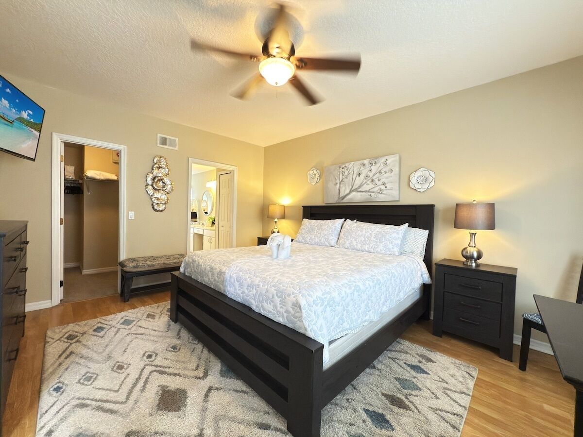 A bedroom with a bed, bedside tables, a ceiling fan, and a view of the hallway.