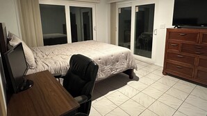 Ein Schlafzimmer mit Bett, Schreibtisch mit Computer, Fernseher auf einer Kommode und einer Glastür, die auf einen Balkon führt.