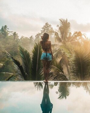 Uma pessoa à beira de uma piscina infinita que reflete a paisagem, com palmeiras ao fundo.