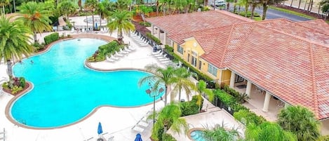 A swimming pool with lounge chairs and umbrellas, surrounded by palm trees and a building with a red-tiled roof.