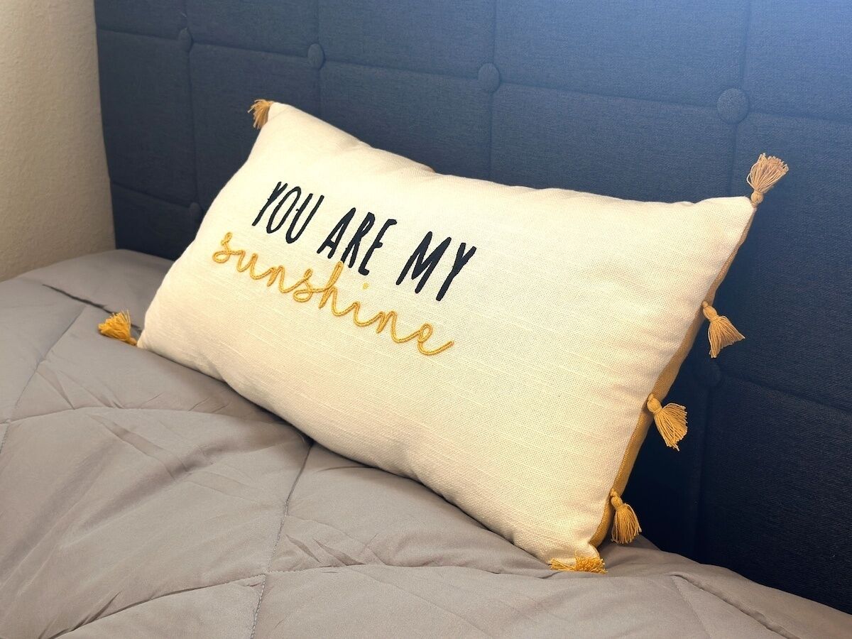 A pillow with embroidered text on a bed with a headboard.