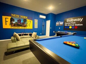 A pool table in a room with a blue wall, a sofa, and a sign for Subway Central Park 5th Avenue Station. - Villa Near Disney Themed Luxury 5bd With Pool 5 Bedroom Home (Kissimmee)