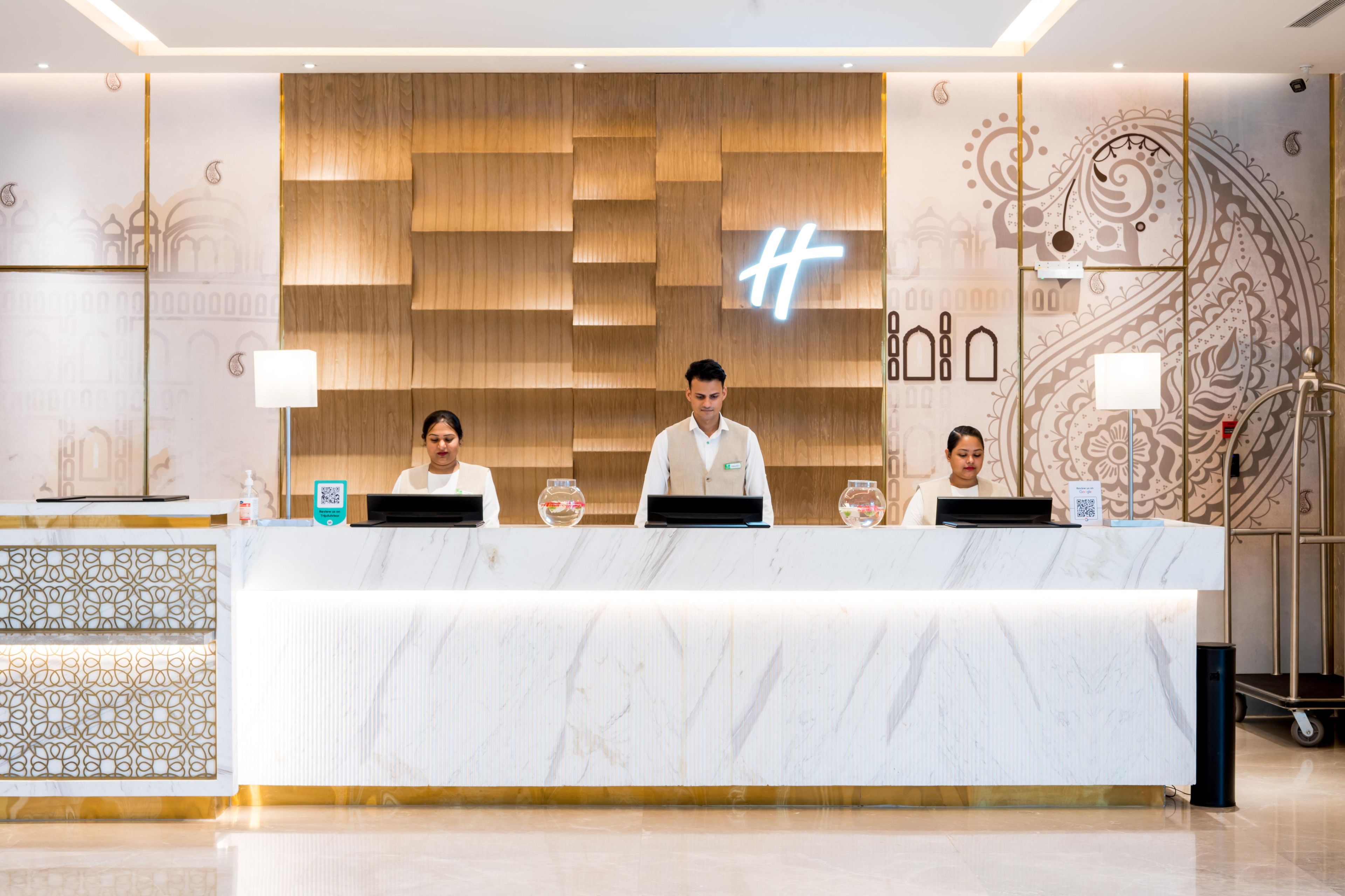 A reception desk with three staff members, a wooden wall with a large 'H' logo, and a patterned wall design.