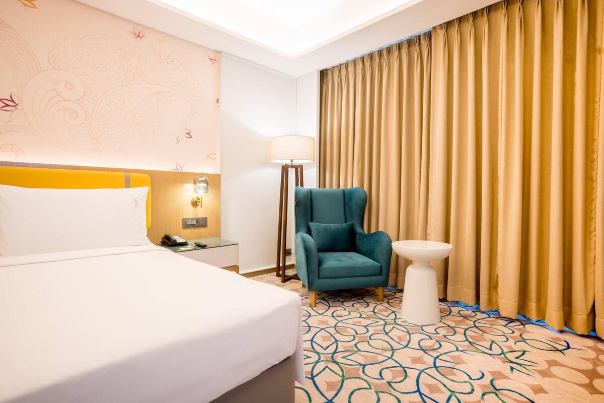 A hotel room with a bed, a chair, a bedside table, a lamp, and a curtain.
