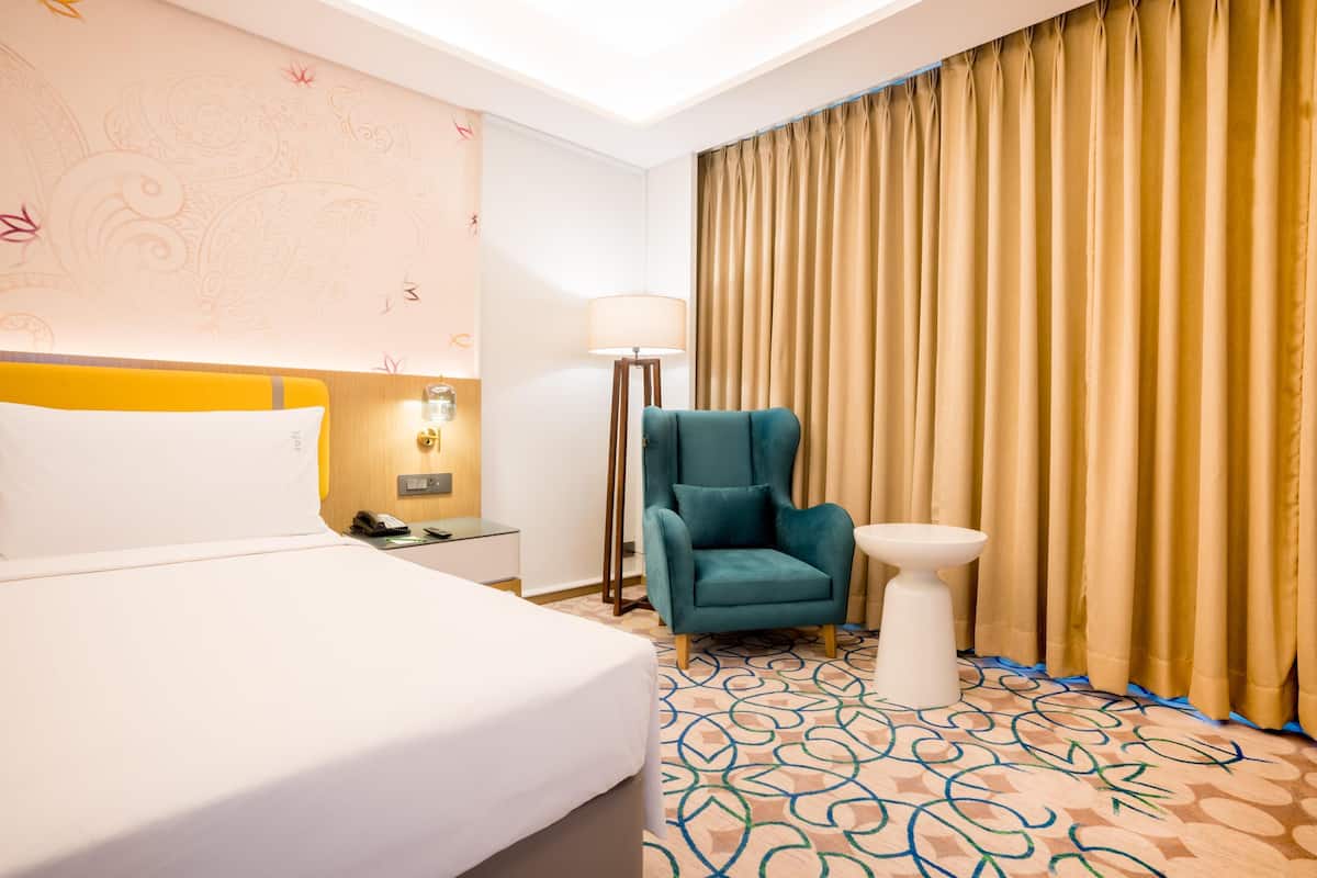 Ein Hotelzimmer mit Bett, Sessel, Nachttisch, Lampe und Vorhang.