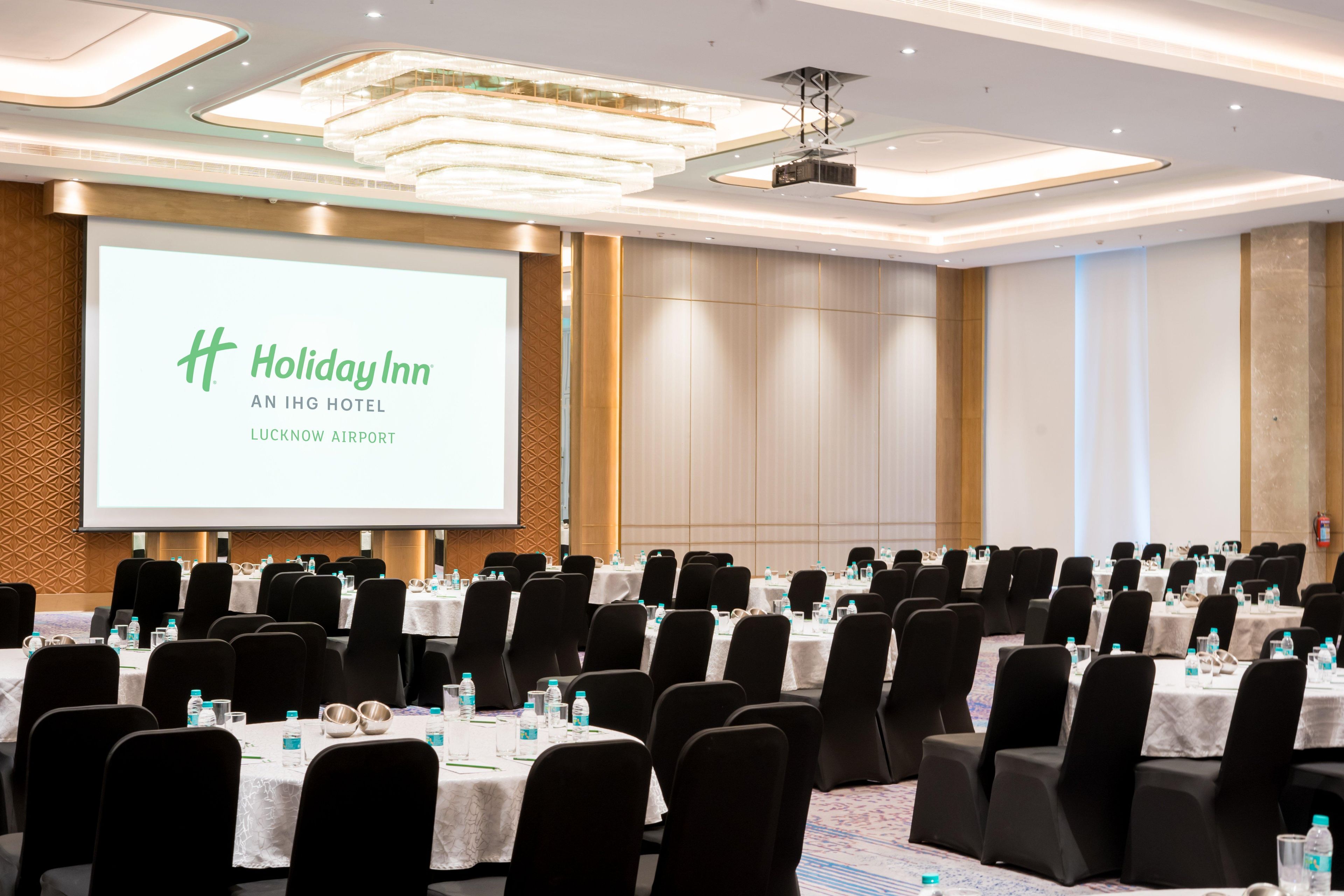 Een congrescentrum met rijen tafels en stoelen, een groot scherm waarop de branding 'Holiday Inn An IHG Hotel Lucknow Airport' staat en een modern plafondontwerp.
