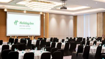 Een congrescentrum met rijen tafels en stoelen, een groot scherm waarop de branding 'Holiday Inn An IHG Hotel Lucknow Airport' staat en een modern plafondontwerp.