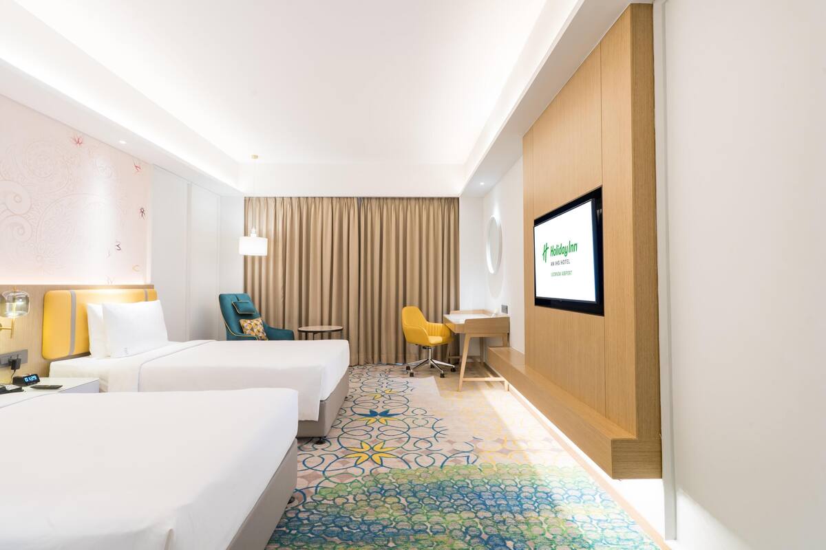 A hotel room with two beds, a desk, a chair, a TV, and a patterned carpet.