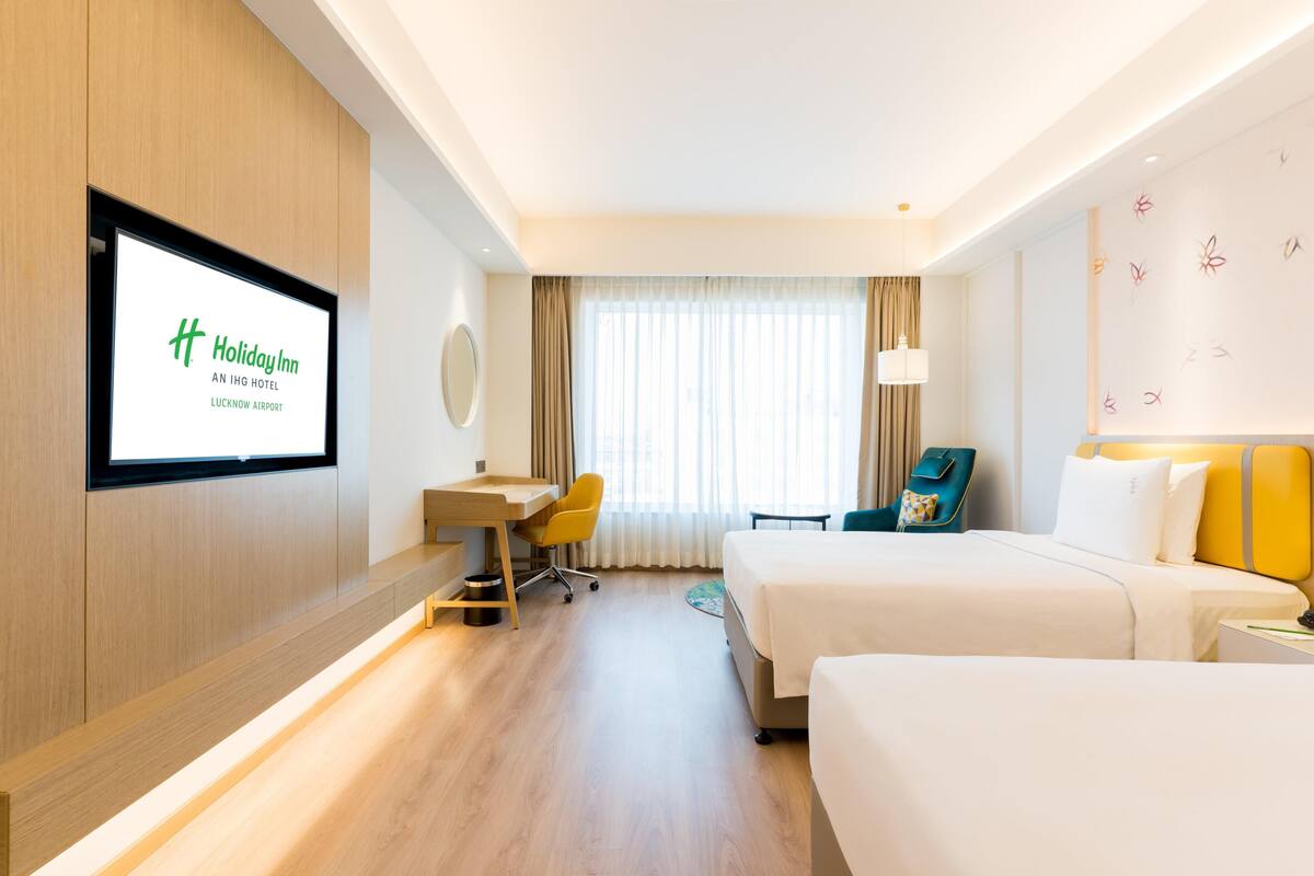 A hotel room with a desk, two beds, a chair, a TV, and a window with curtains.