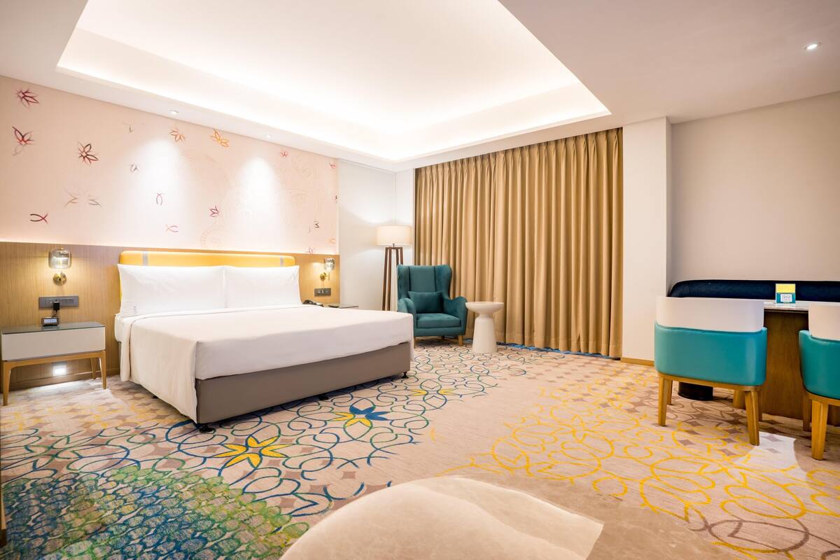 A hotel room with a bed, a desk, a chair, a lamp, and a patterned carpet.