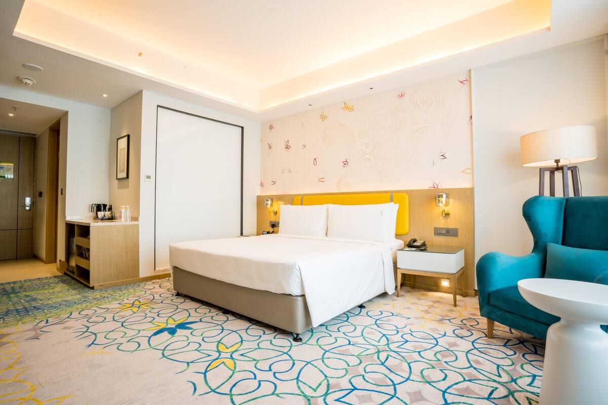 A hotel room with a bed, bedside table, lamp, chair, and a desk with a lamp.