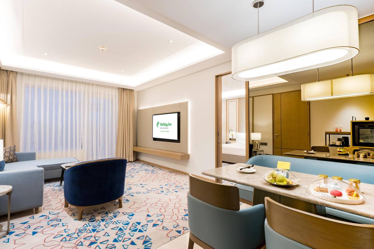 A hotel room with a seating area, a dining table with a breakfast tray, a large mirror, and a screen displaying hotel information.